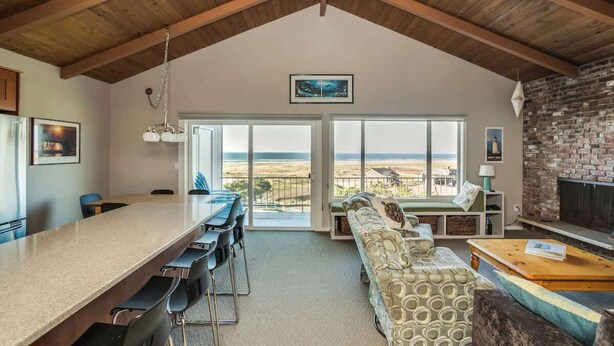 A spacious living area with a high ceiling, a long dining table, a sofa, and a fireplace. - Pacific View V486 (Gearhart)