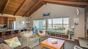 A spacious living area with a high ceiling, a kitchen with modern appliances, and a large window offering a view of the landscape. - Pacific View V486 (Gearhart)