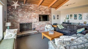 A living room with a brick fireplace, wooden ceiling beams, a television, and a sofa. - Pacific View V486 (Gearhart)