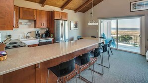A kitchen with wooden cabinets, a stainless steel refrigerator, a large island with bar stools, and a view of the beach through the sliding glass door. - Pacific View V486 (Gearhart)