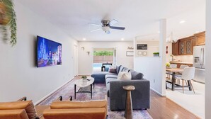 A modern living room with a sofa, coffee table, and chairs. There is a dining area with a table and chairs. A kitchen is visible with cabinets and appliances.