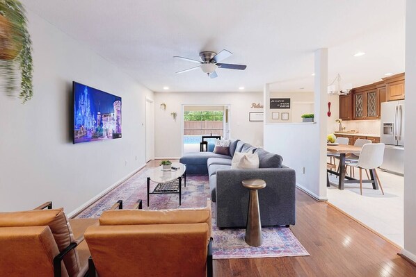 A modern living room with a sofa, coffee table, and chairs. There is a dining area with a table and chairs. A kitchen is visible with cabinets and appliances.