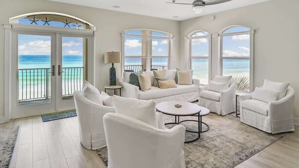 A spacious living room with a white sofa set, a coffee table, and a ceiling fan. Large windows offer a view of the ocean.