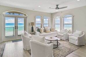 Un salon spacieux avec un ensemble de canapés blancs, une table basse et un ventilateur de plafond. De grandes fenêtres offrent une vue sur l’océan.