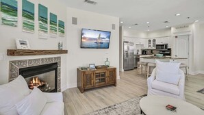 A modern living room with a fireplace, white furniture, a flat-screen TV, and a kitchen in the background.