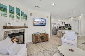 A modern living room with a fireplace, white furniture, a flat-screen TV, and a kitchen in the background. - Sunset Villa - Unit C-302 (Santa Rosa Beach)