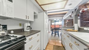 Uma cozinha moderna com armários brancos, fogão e geladeira. Uma área de refeições com uma mesa e cadeiras fica visível por uma porta aberta.