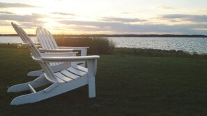 Två vita Adirondack-stolar placerade framför en sjö eller ett annat vattendrag under solnedgången.