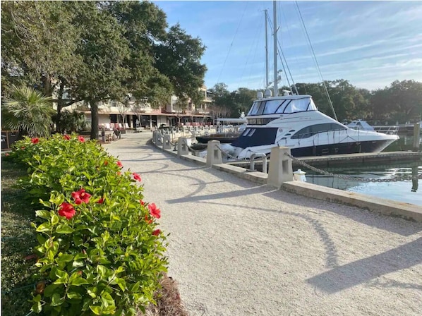 A marina with a docked yacht, a pathway lined with greenery, and a building in the background.