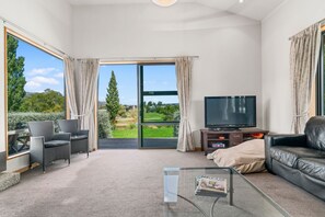 A spacious living room with a large sliding glass door leading to a garden, a flat-screen TV, a sofa, and a coffee table.