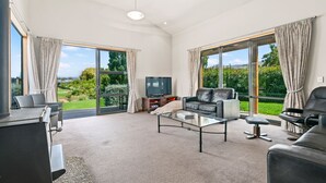 A spacious living room with a large TV, comfortable seating, and large windows offering a view of greenery. - Edgewater Escape - Cromwell Holiday Home (Cromwell)