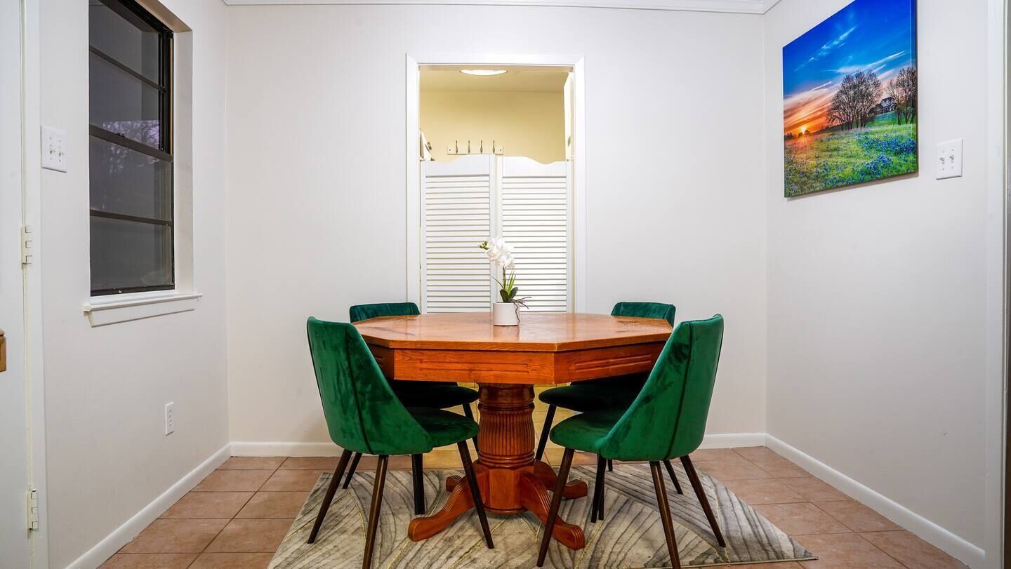 Un comedor con una mesa redonda de madera, sillas tapizadas de verde, una ventana con persianas y un cuadro en la pared.