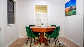 Un comedor con una mesa redonda de madera, sillas tapizadas de verde, una ventana con persianas y un cuadro en la pared.