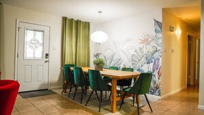 A dining area with a wooden table, green chairs, a potted plant, and a wall mural.