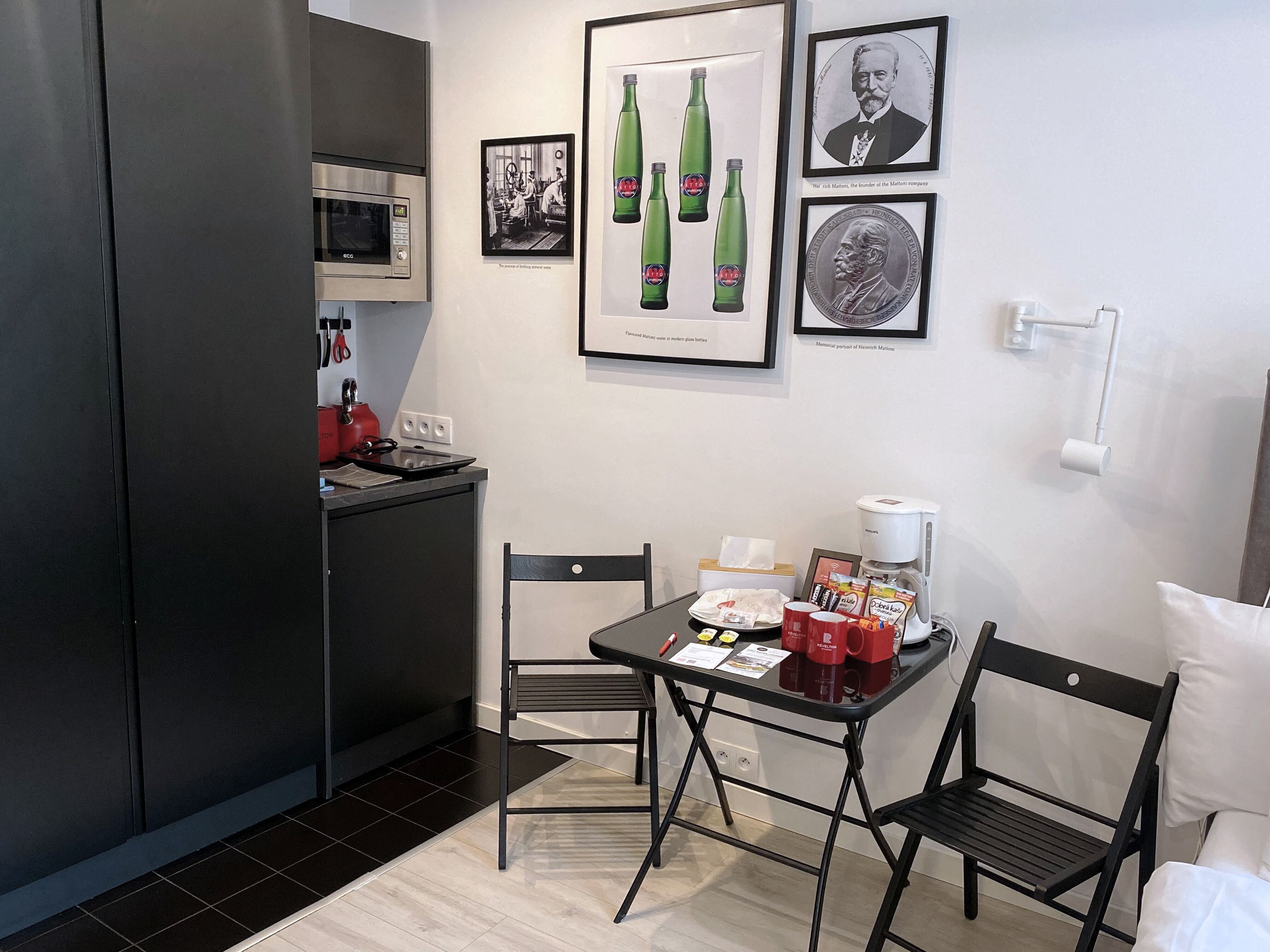 Eine kompakte Küche mit Mikrowelle, Herd und Kaffeemaschine. An der Wand befinden sich gerahmte Bilder, darunter ein Porträt und eine alte Werbeanzeige. Im Essbereich stehen zwei Stühle und ein kleiner Tisch.