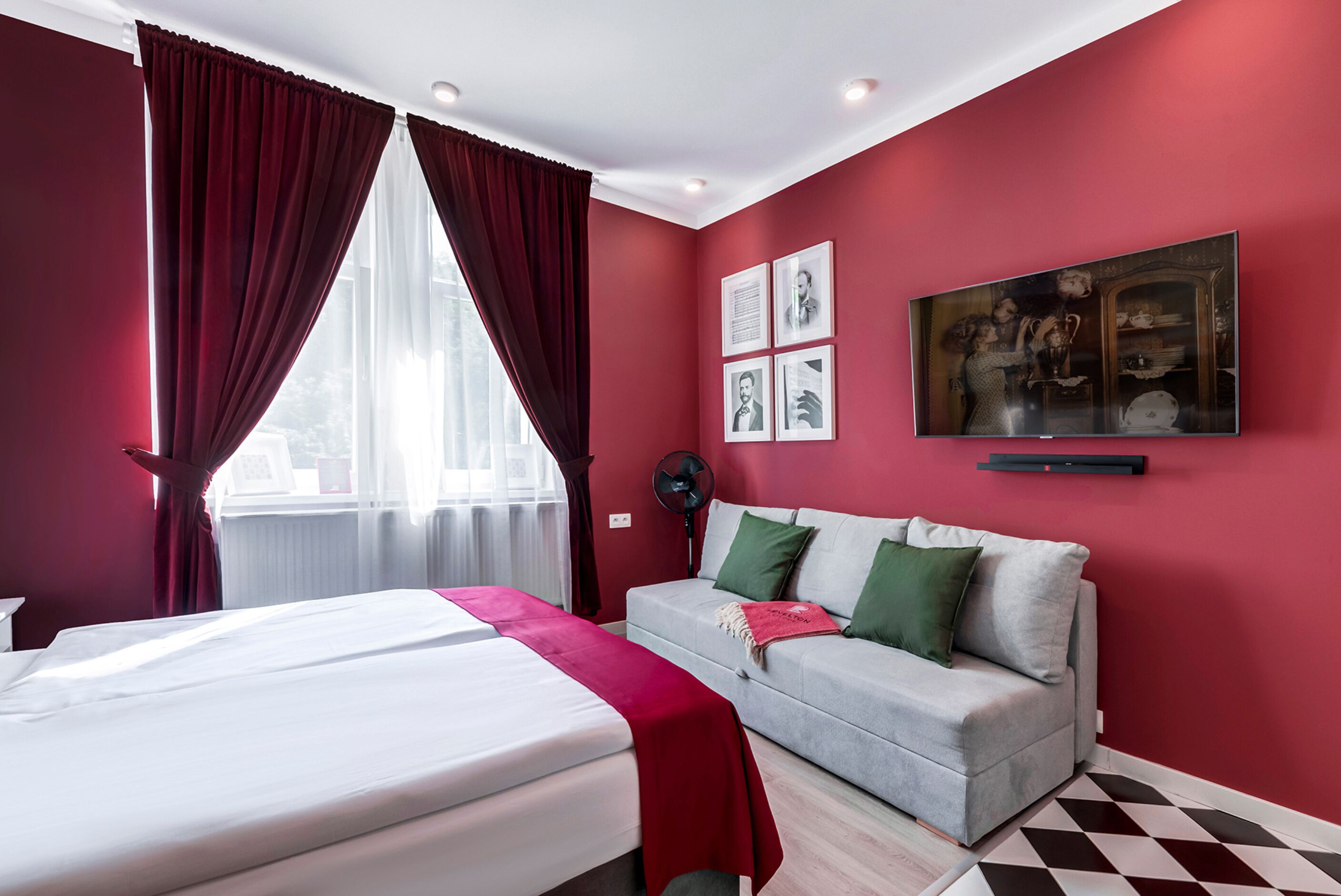 Una habitación de hotel con cama, sofá, cortinas rojas, ventilador y obras de arte en la pared.