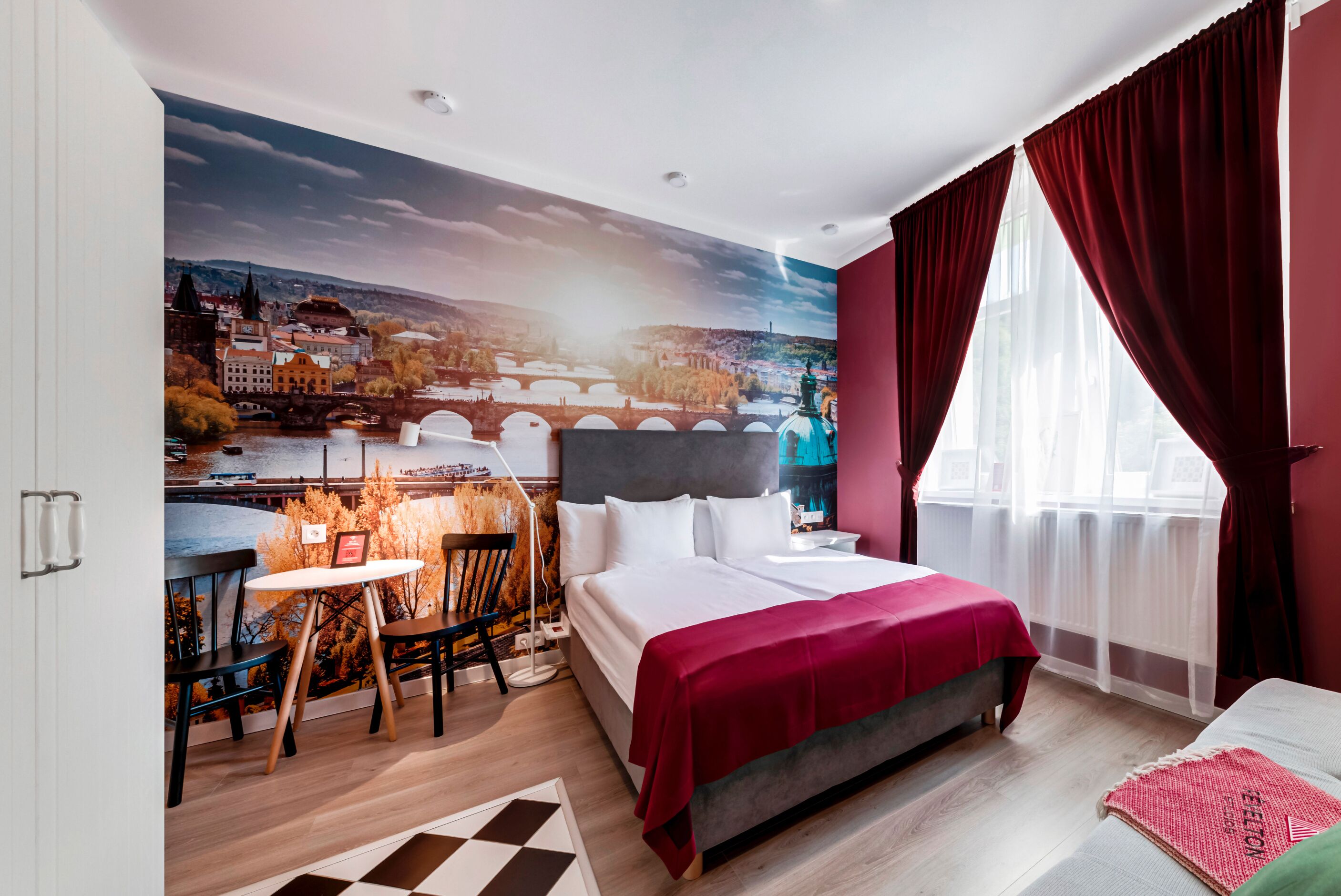 A hotel room with a large mural of a cityscape, a bed with a red blanket, a desk with a chair, and a window with red curtains.