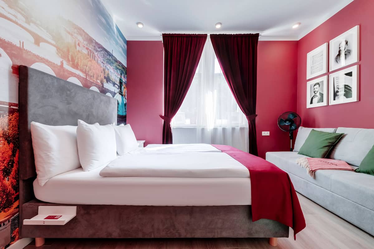 Moderni hotellihuone, jossa on suuri sänky, harmaa sohva, punaiset verhot ja seinällä kaupunkimaisemaa esittävä seinämaalaus.
