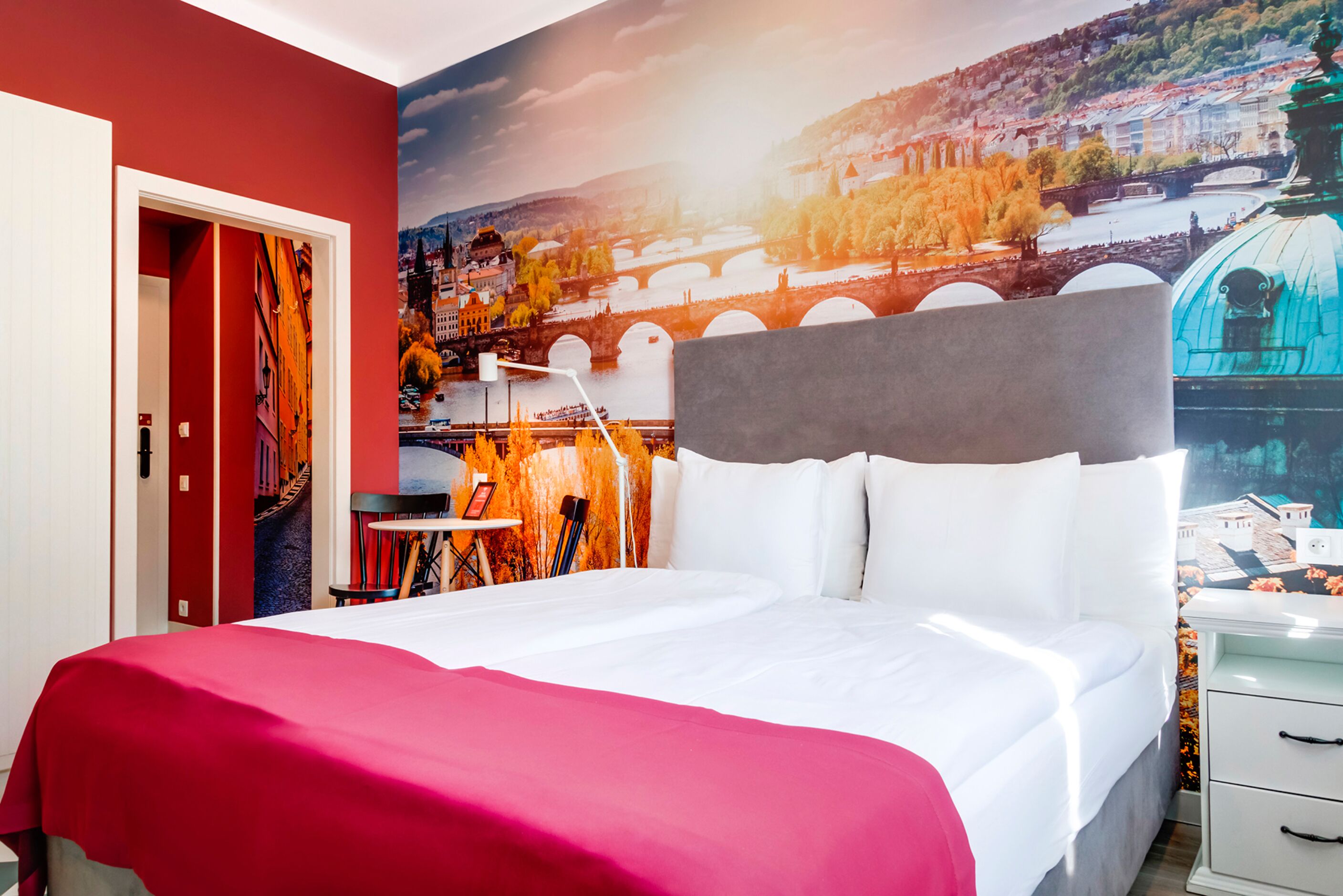 Habitación de hotel con una cama grande, una pared roja y un mural de un paisaje urbano.
