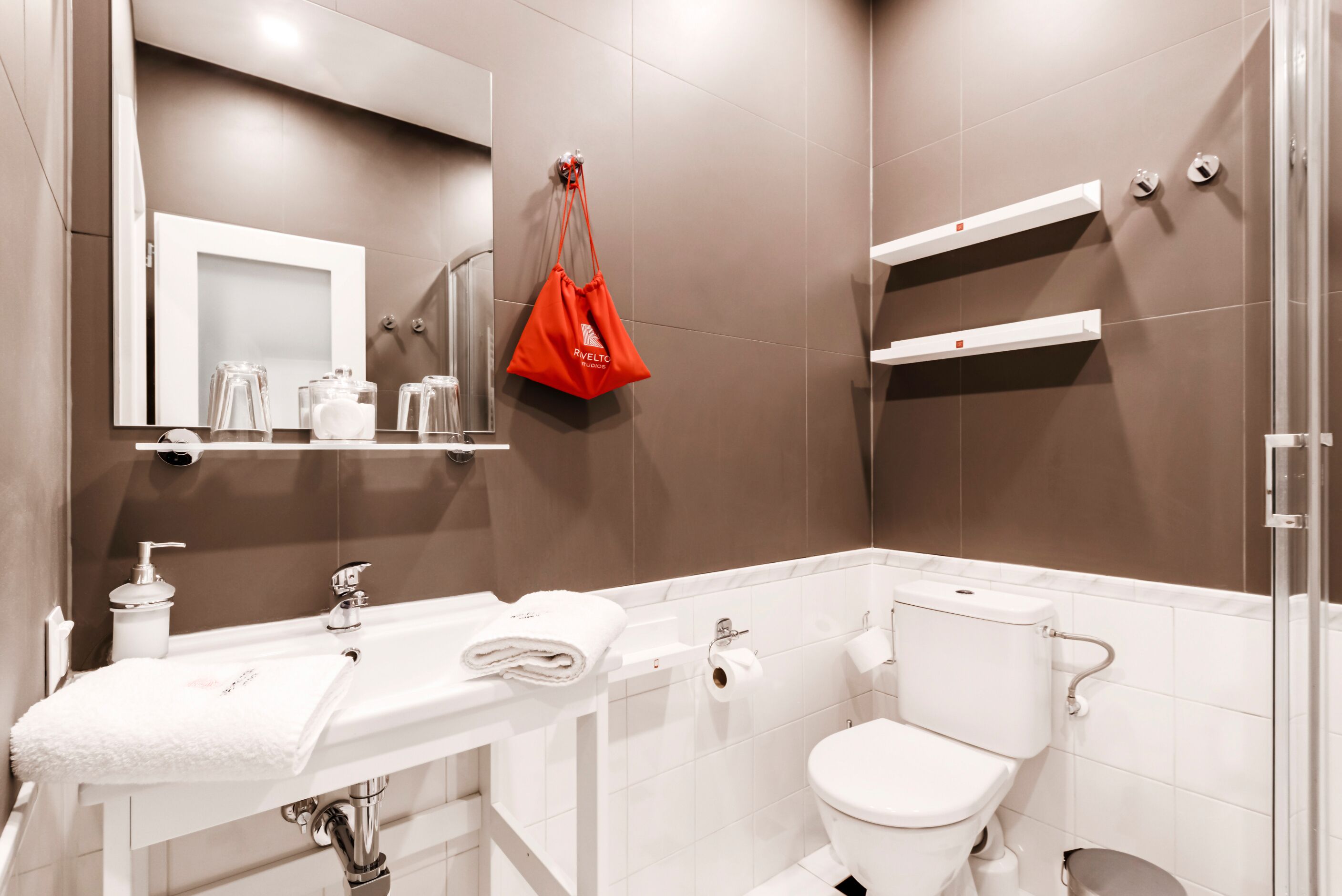 A modern bathroom with a white sink, mirror, and toilet. There is a red bag hanging on the wall and a shelf with toiletries.