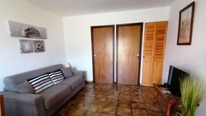 Una sala de estar con un sofá gris, un televisor, una puerta de madera y piso de baldosas.