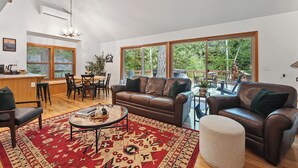 A spacious living room with a large window, a brown leather sofa, a coffee table, and a red patterned rug. - Guest Favorite Mt. Hood Cabin - Private Hot Tub - Sandy River (Rhododendron)