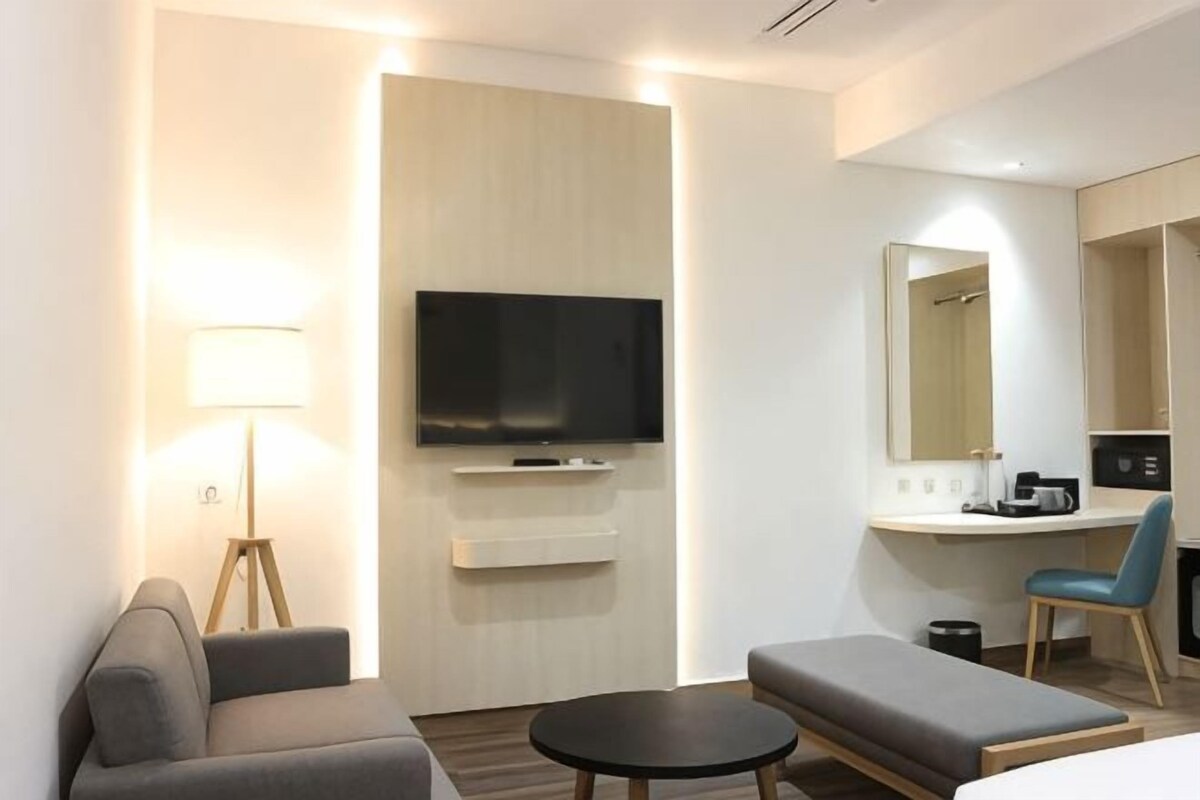 Una habitación de hotel moderna con una televisión de pantalla plana, un sofá, una pequeña mesa redonda, una silla, un escritorio con una computadora y un espejo.
