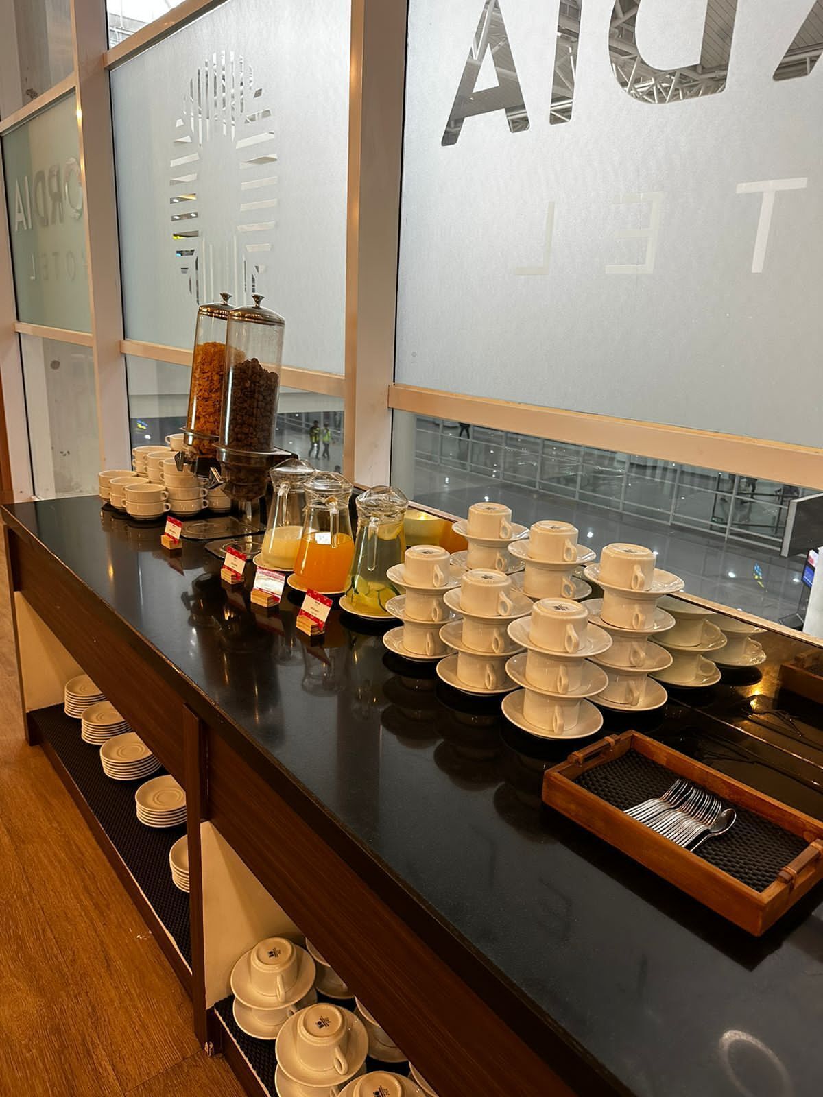 A breakfast buffet with coffee, tea, and juice dispensers, neatly arranged white cups and saucers, and a tray of utensils.