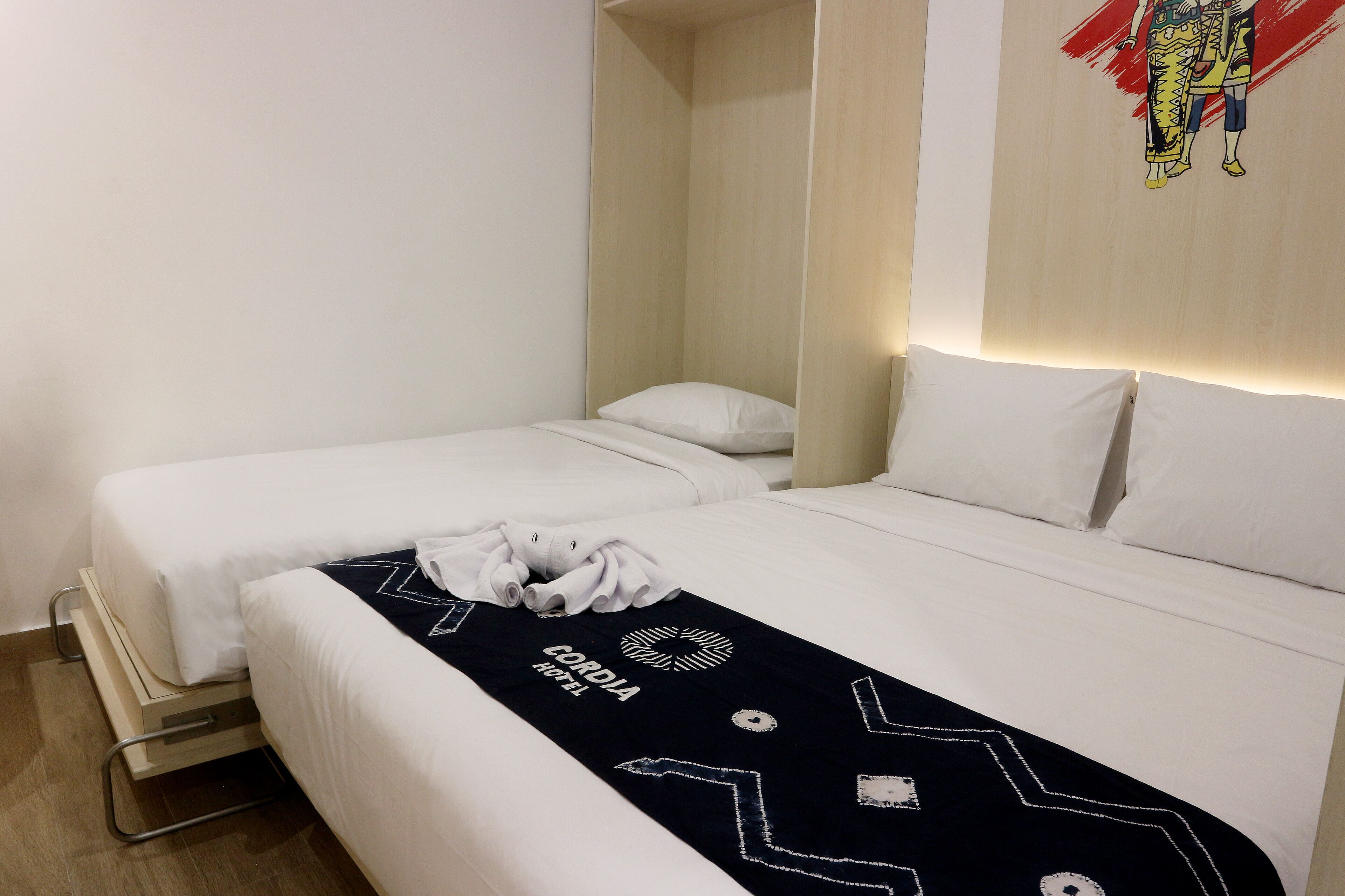 Habitación de hotel con dos camas individuales, un corredor de cama blanco y negro con el nombre del hotel, una toalla blanca en el suelo y una obra de arte colgada en la pared.