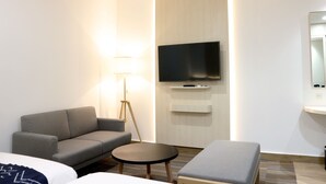 A modern hotel room with a grey sofa, a small round table, a flat-screen TV mounted on the wall, and a bedside table. - Cordia Hotel Banjarmasin - Hotel Dalam Bandara (Banjarbaru)