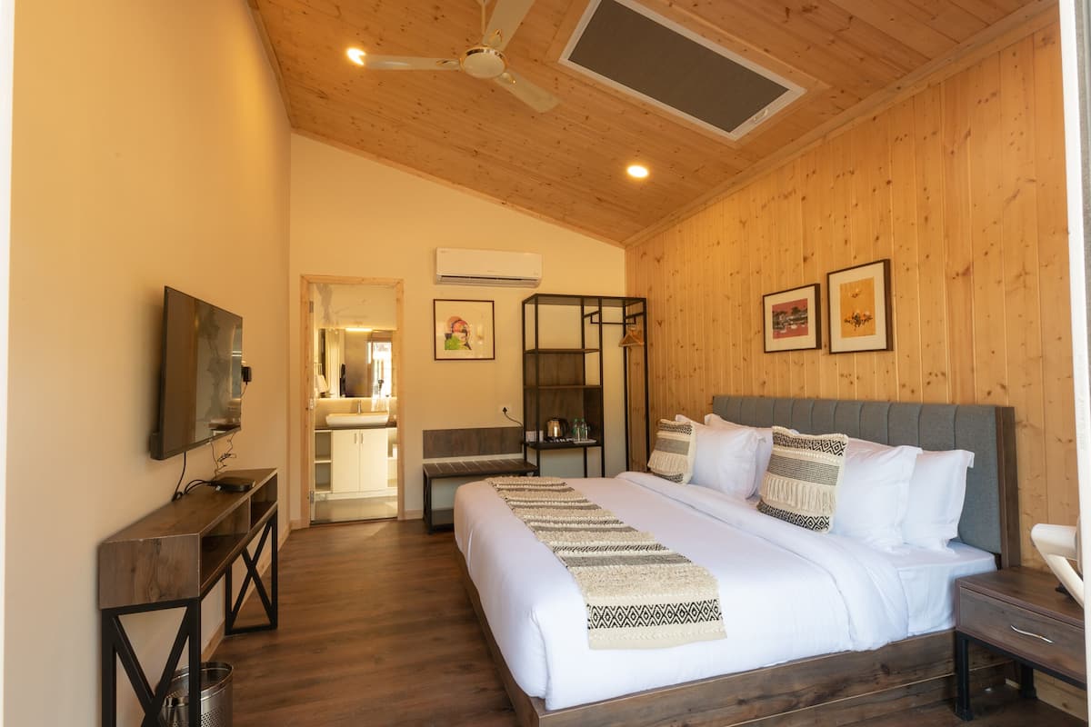 Een slaapkamer met een houten plafond, een bed met wit beddengoed, een nachtkastje, een televisie op een standaard en een badkamer zichtbaar door een geopende deur.