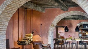 Un intérieur rustique avec des arches en briques apparentes, une cheminée, un coin repas avec une table et des chaises, et une lampe suspendue.
