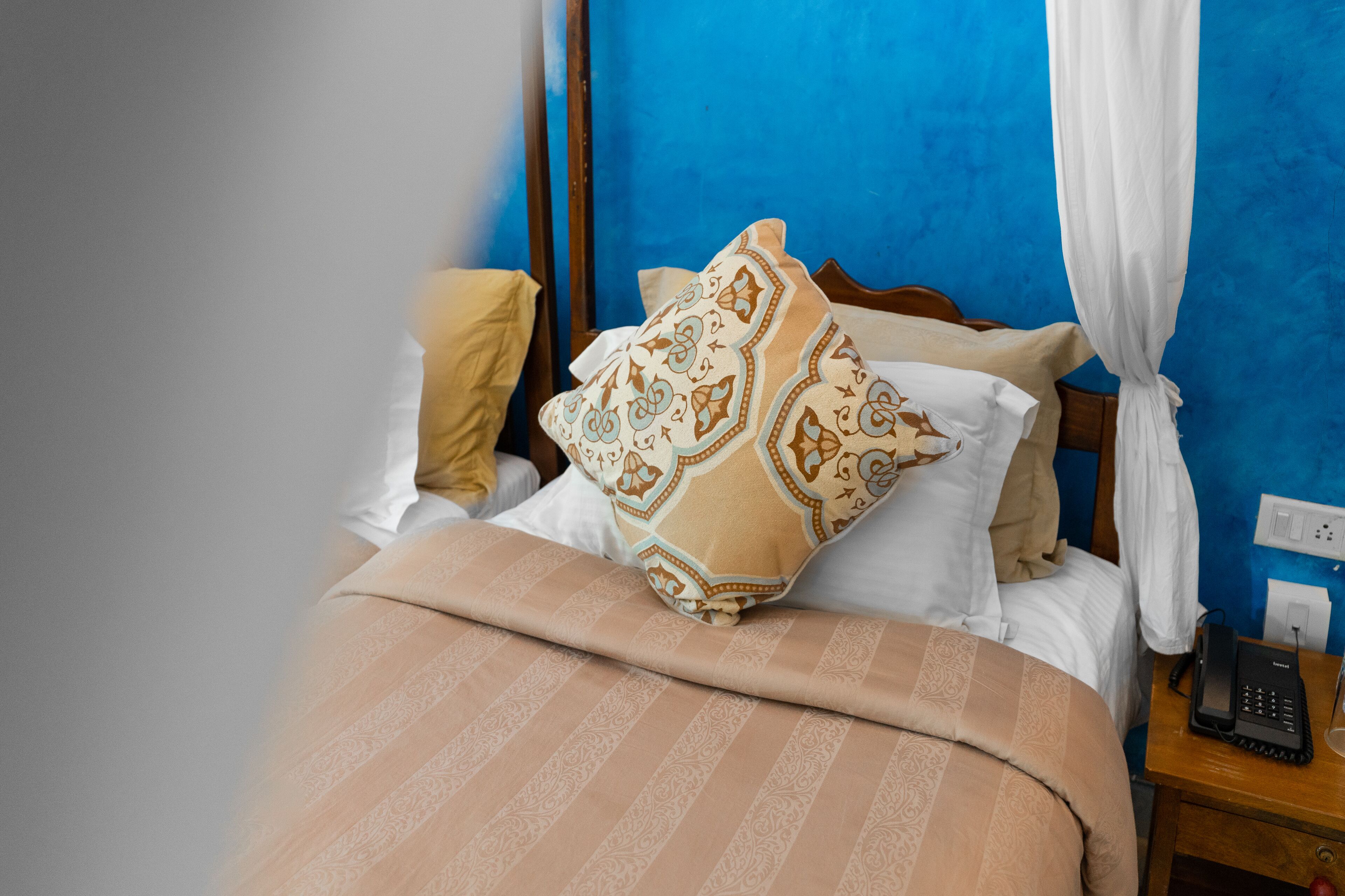 A neatly made bed with patterned pillows and a striped blanket, a bedside telephone, and a power outlet on a wooden nightstand.