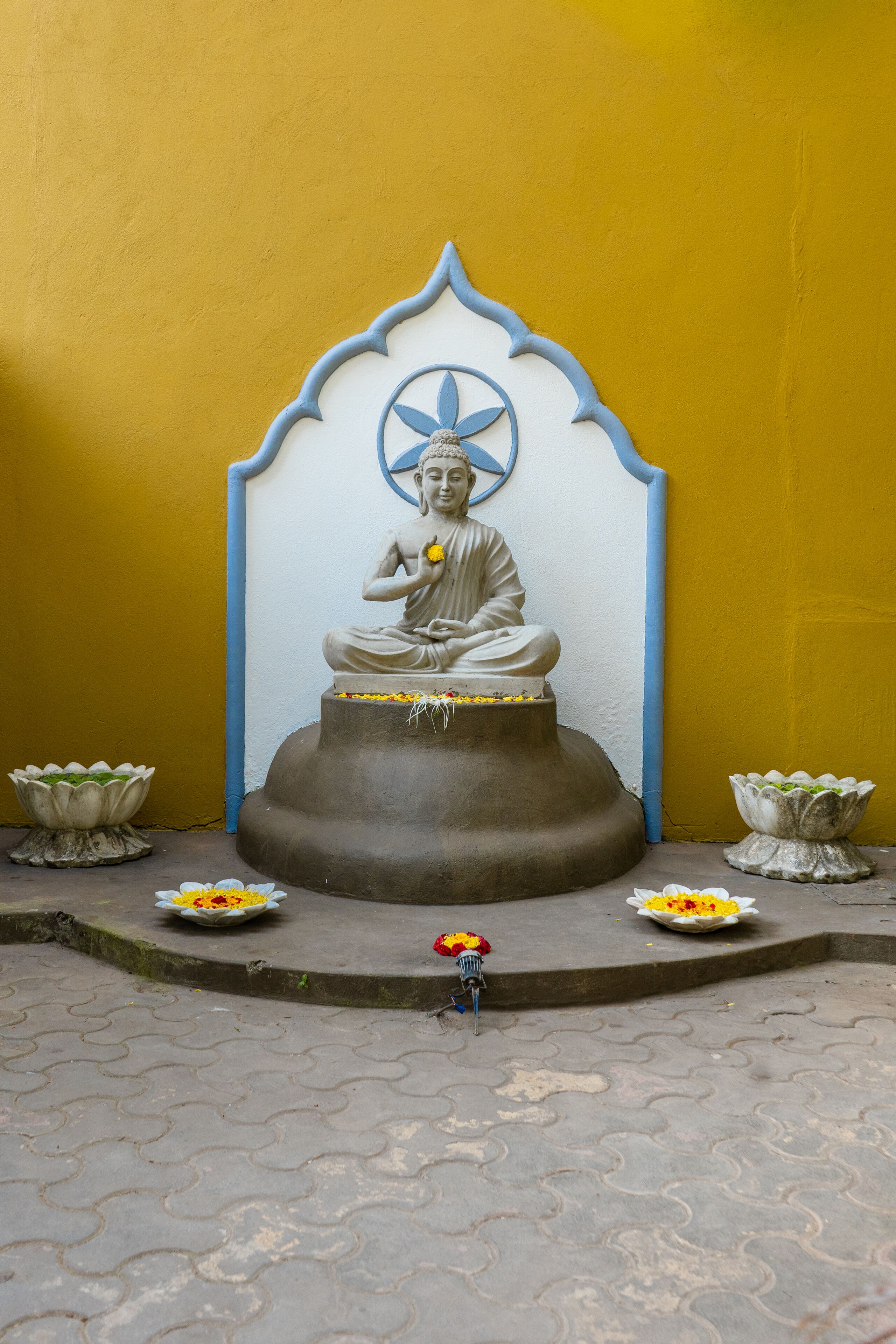 A statue of a seated figure with a blue halo, surrounded by flower offerings on a tiled floor.