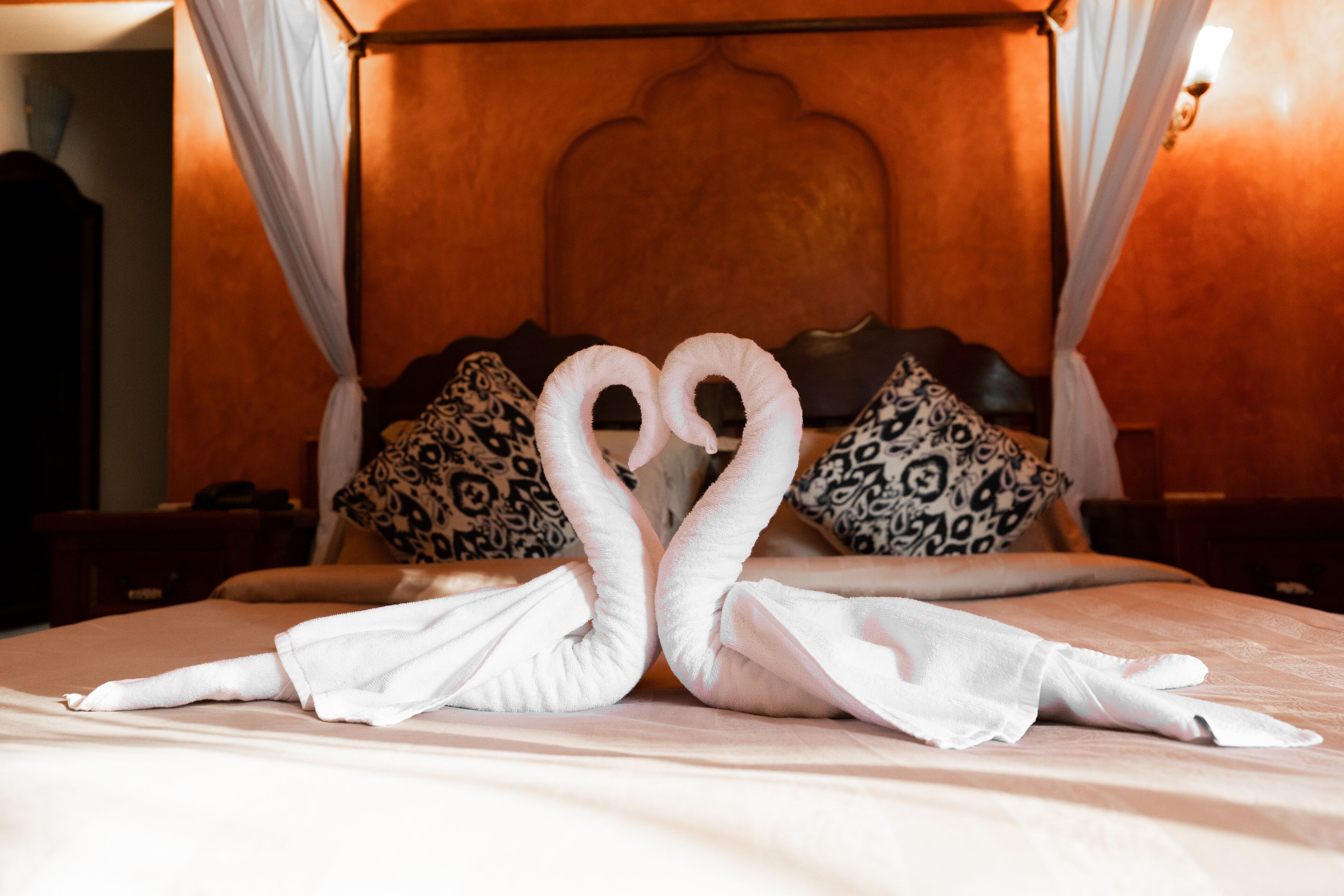 A bed with two towels arranged to resemble swans, a wooden headboard, patterned pillows, and a nightstand.