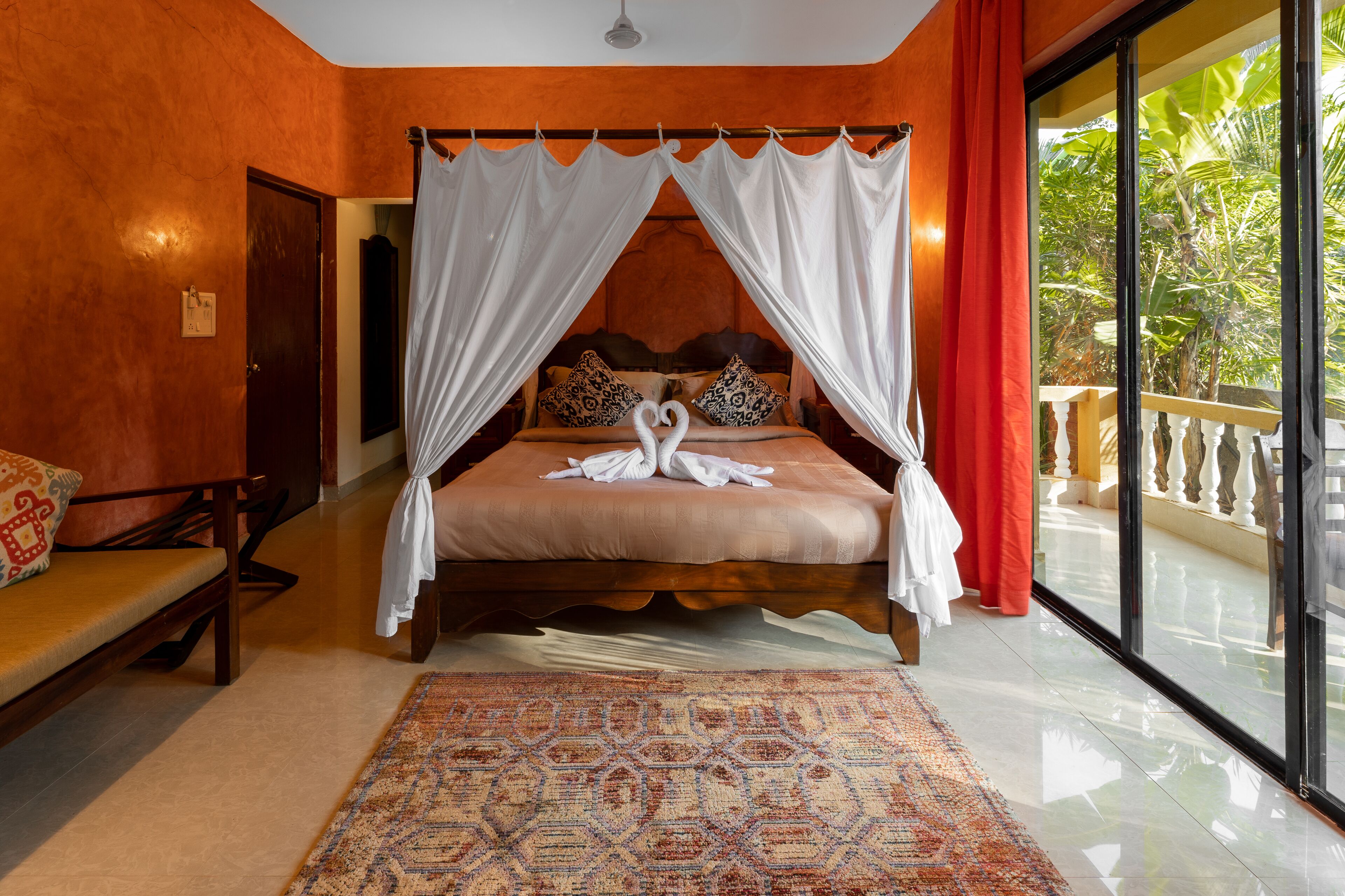 A bedroom with a four-poster bed, a large window with a view of greenery, a patterned rug, and a wooden bench.
