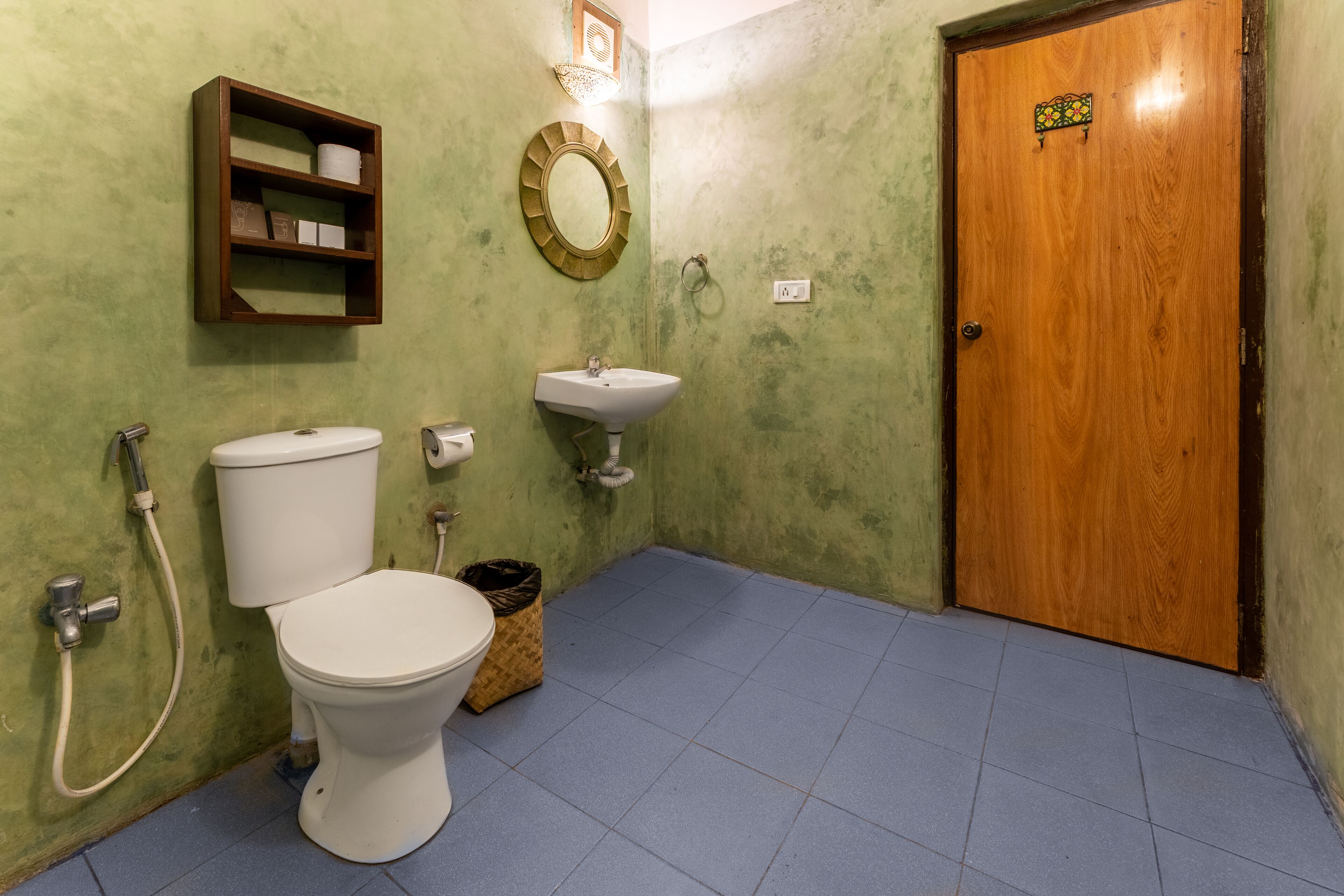 Casa de banho com sanita, lavatório e chuveiro. Tem uma porta de madeira e uma prateleira com artigos de higiene pessoal.
