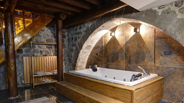 Una bañera de madera en un cuarto con paredes de piedra, una escalera de madera y un estante con toallas.