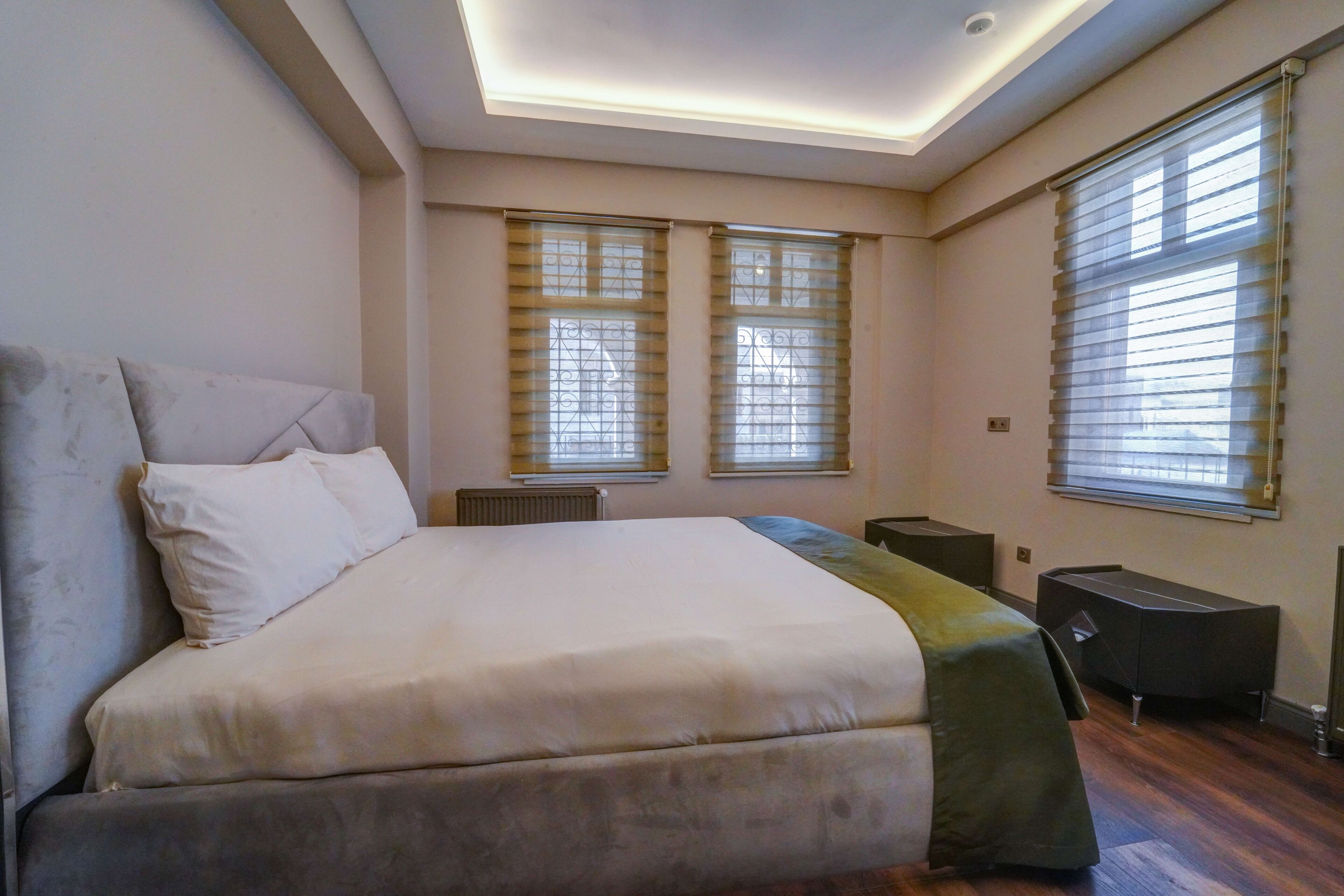 Quarto com cama grande, duas janelas com persianas, escrivaninha e radiador.