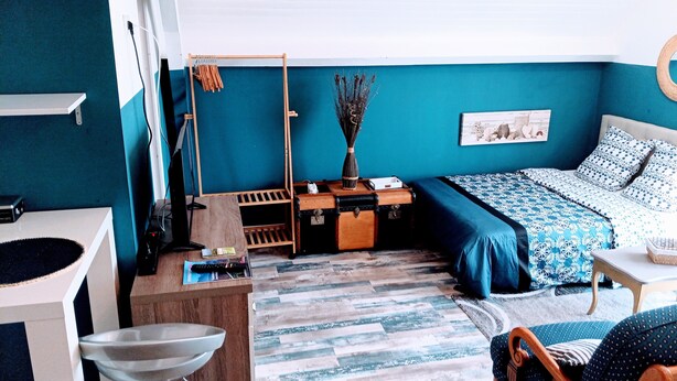 A room with teal walls, a bed with a blue patterned bedspread, a wooden desk with a computer, a chair, and a small table with a vase. - Casa Azur - Charming Palois studio with free parking 24/24 (Pau)
