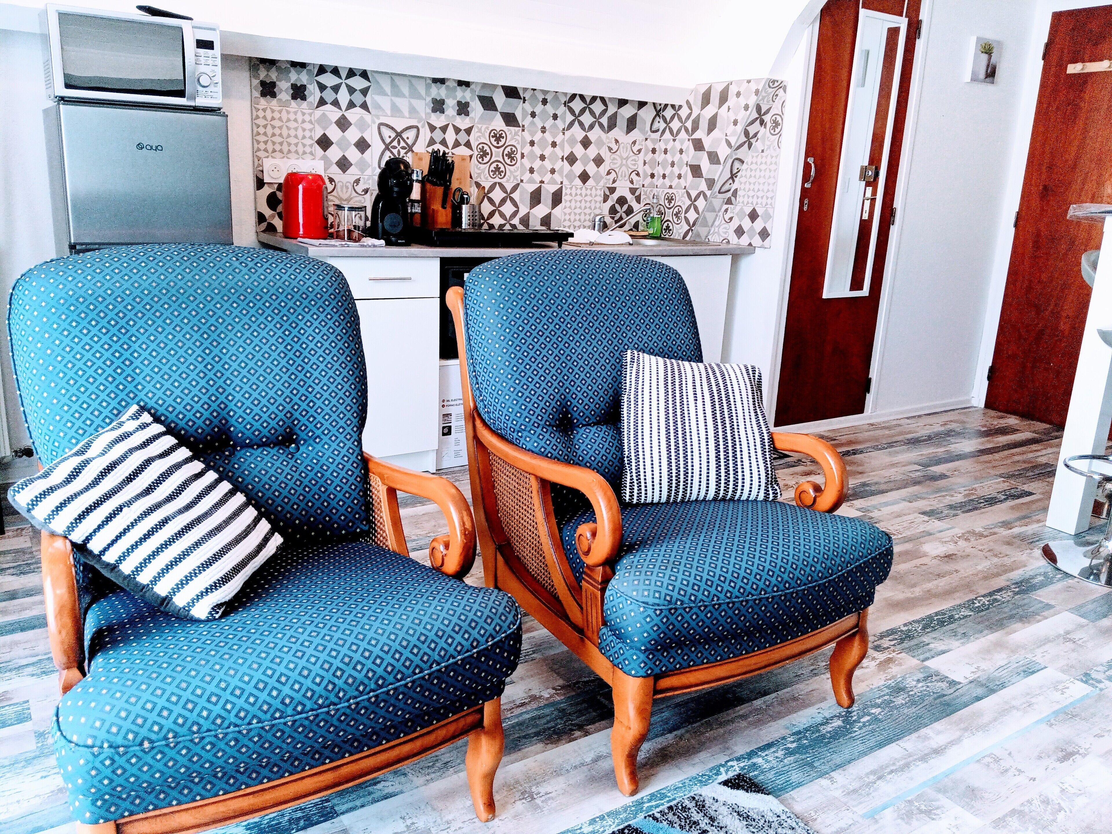 A kitchen with a microwave and refrigerator, two blue patterned chairs with cushions, and a patterned backsplash.