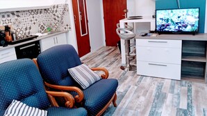 A modern living room with a blue sofa, a wooden chair, a kitchen area with a microwave, and a TV on a stand. - Casa Azur - Charming Palois studio with free parking 24/24 (Pau)
