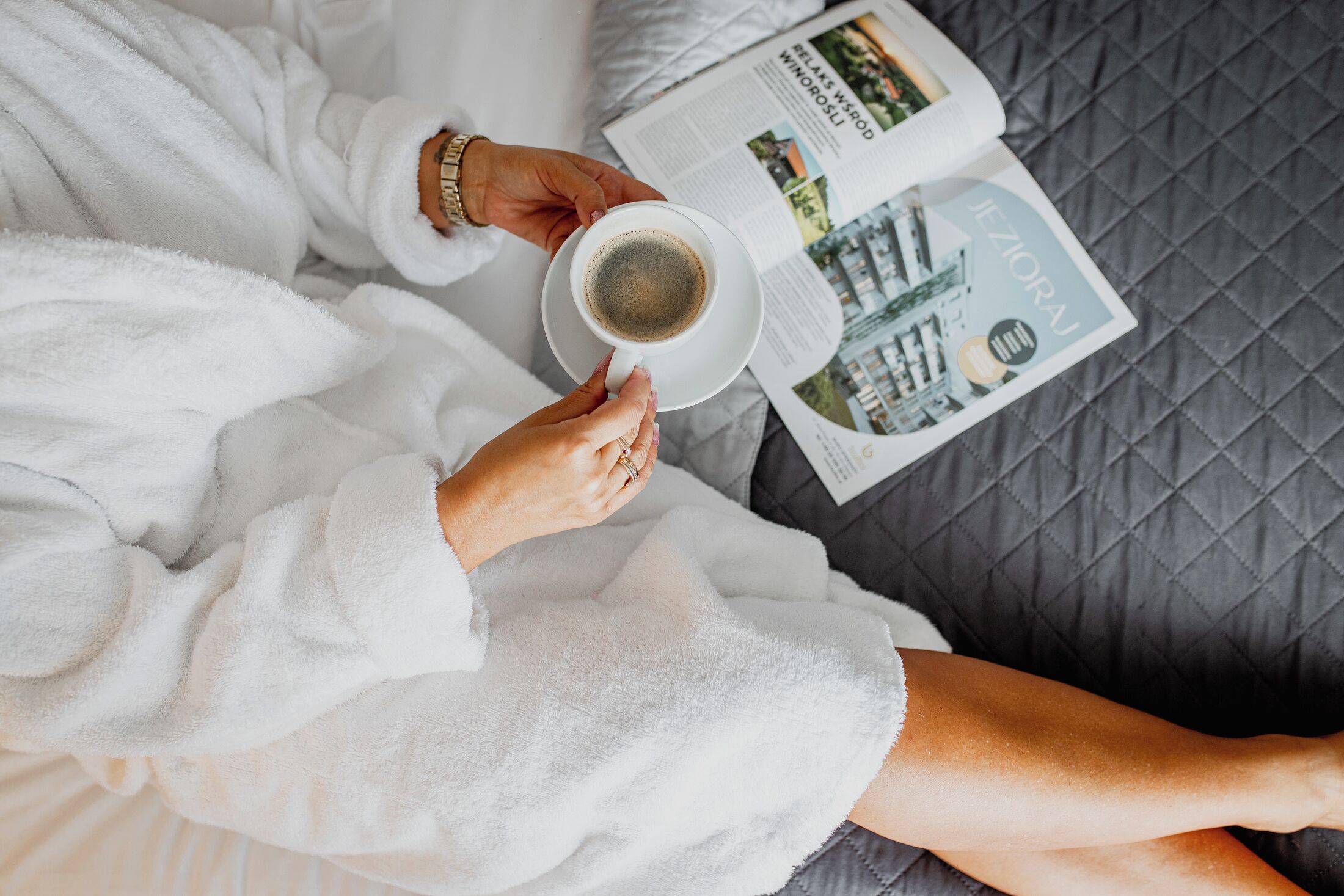 Una persona con bata blanca sostiene una taza de café y una revista sobre una cama con cubierta gris acolchada.