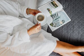 A person in a white bathrobe holding a cup of coffee and a magazine on a bed with a quilted gray cover.