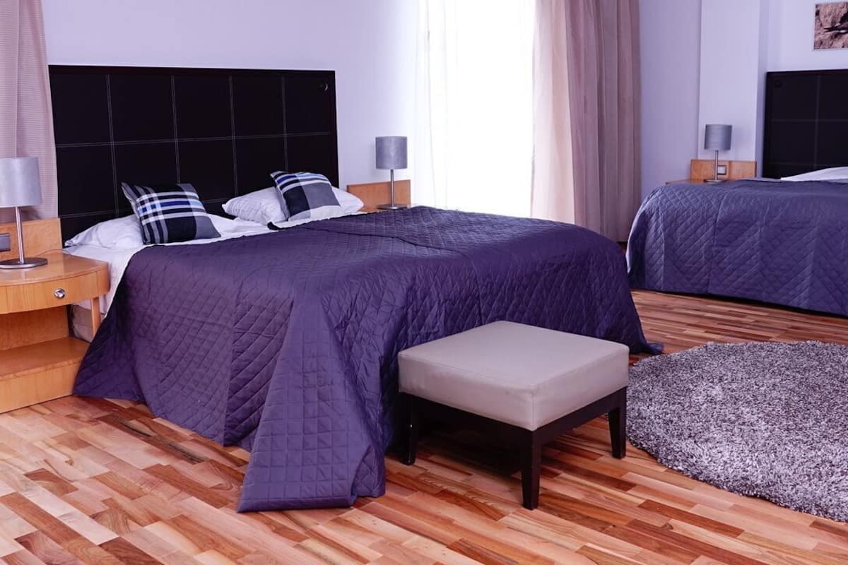 A hotel room with two beds, a bench, a rug, and a nightstand with a lamp.