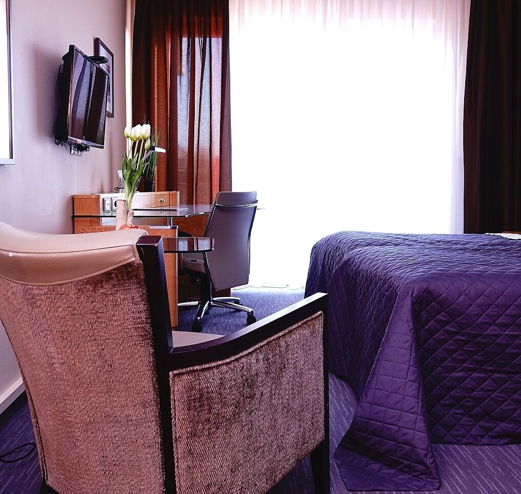 Ein Hotelzimmer mit Bett, Schreibtisch, Stuhl, Fernseher und einem Fenster mit Vorhängen.