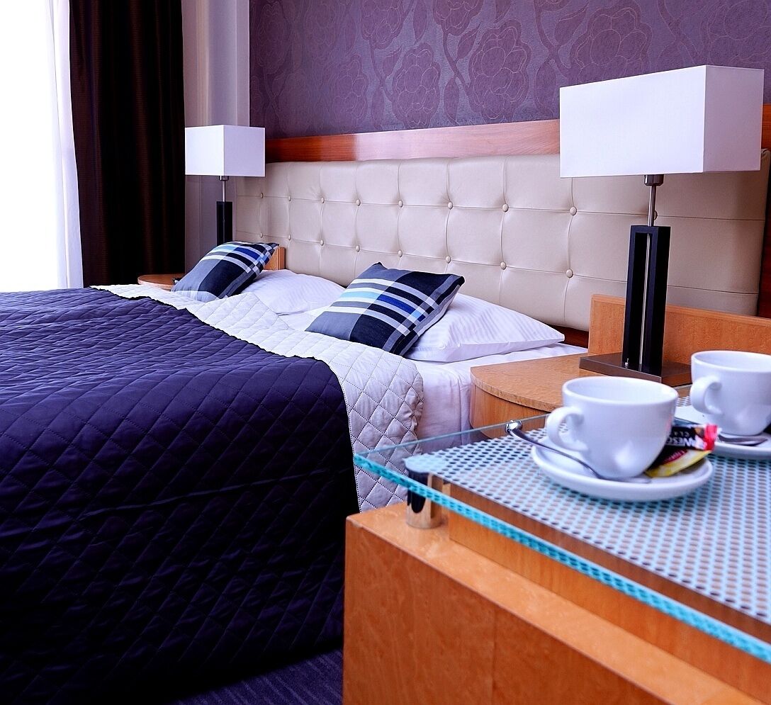 A hotel room with a large bed, two bedside tables, two lamps, and a tray table with cups and a book.