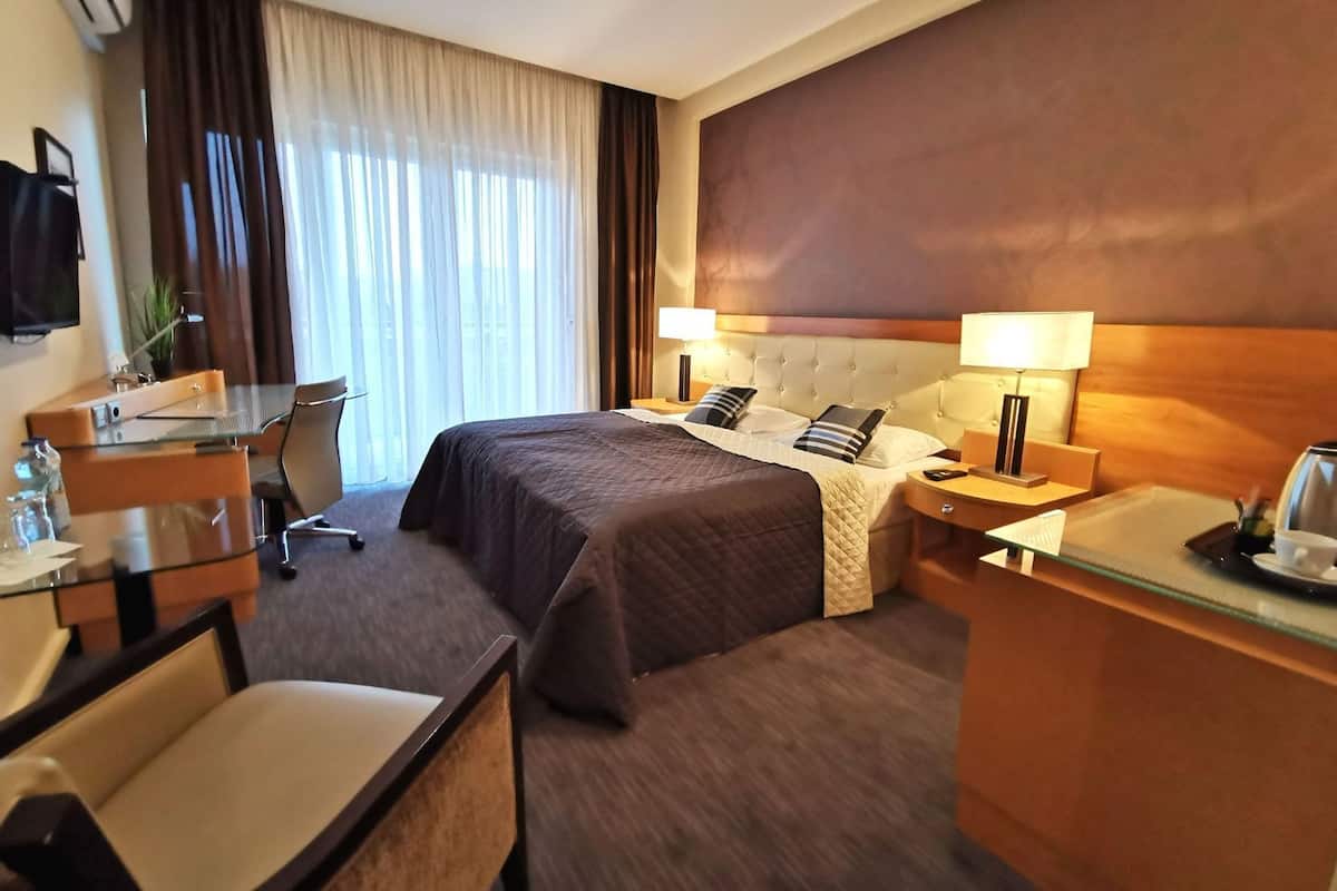 A hotel room with a bed, bedside tables, a desk, a chair, a TV, and a window with curtains.