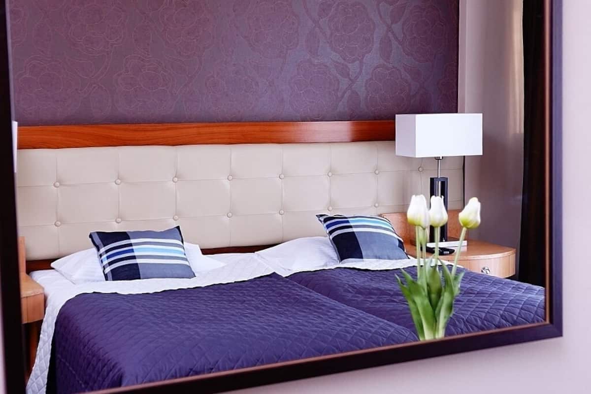 A hotel room with a large bed, two pillows, a nightstand with a lamp, and a mirror reflecting the room.