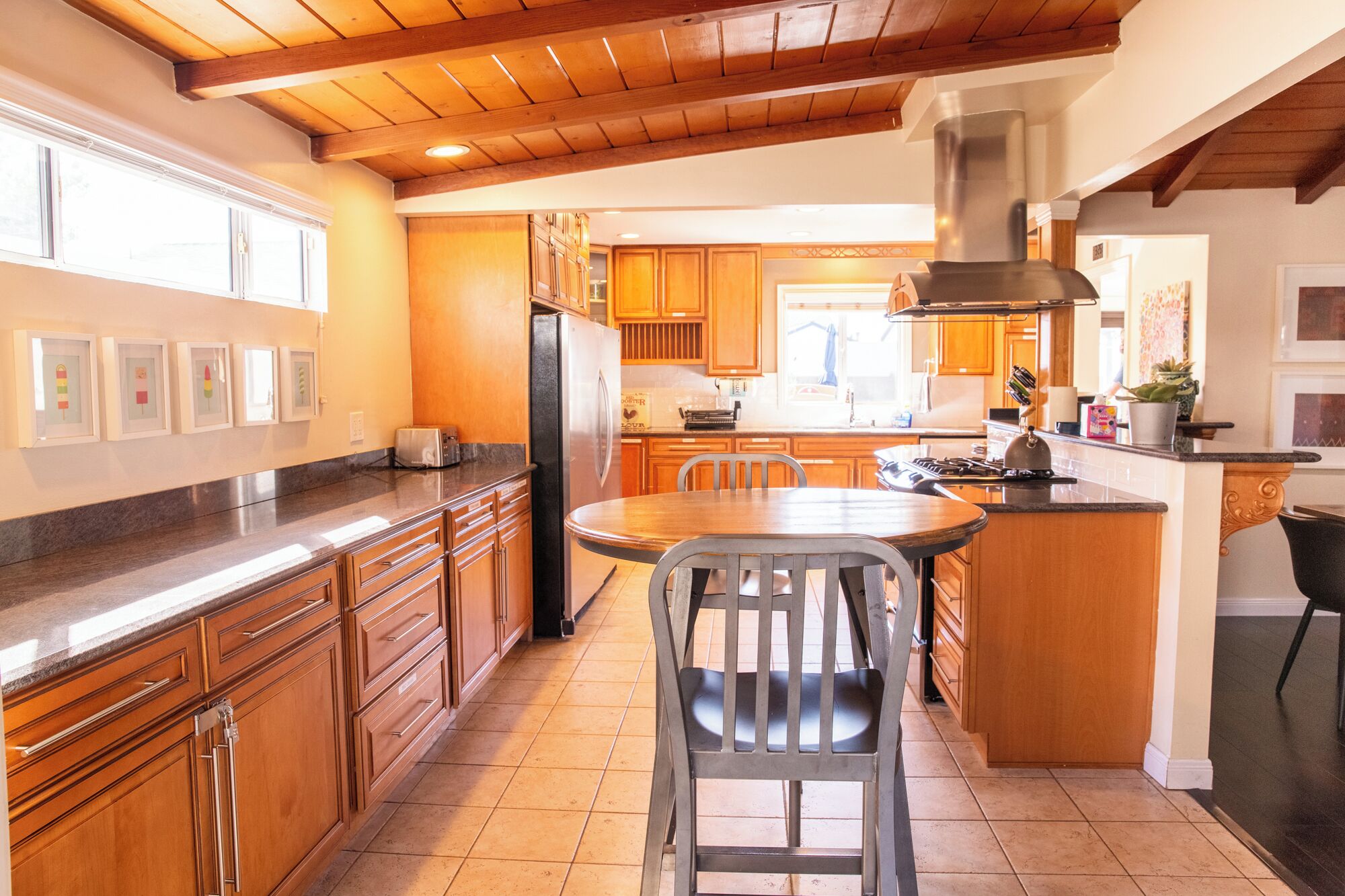A kitchen with wooden cabinets, a stainless steel refrigerator, a dining table with chairs, and a large island with a stove.